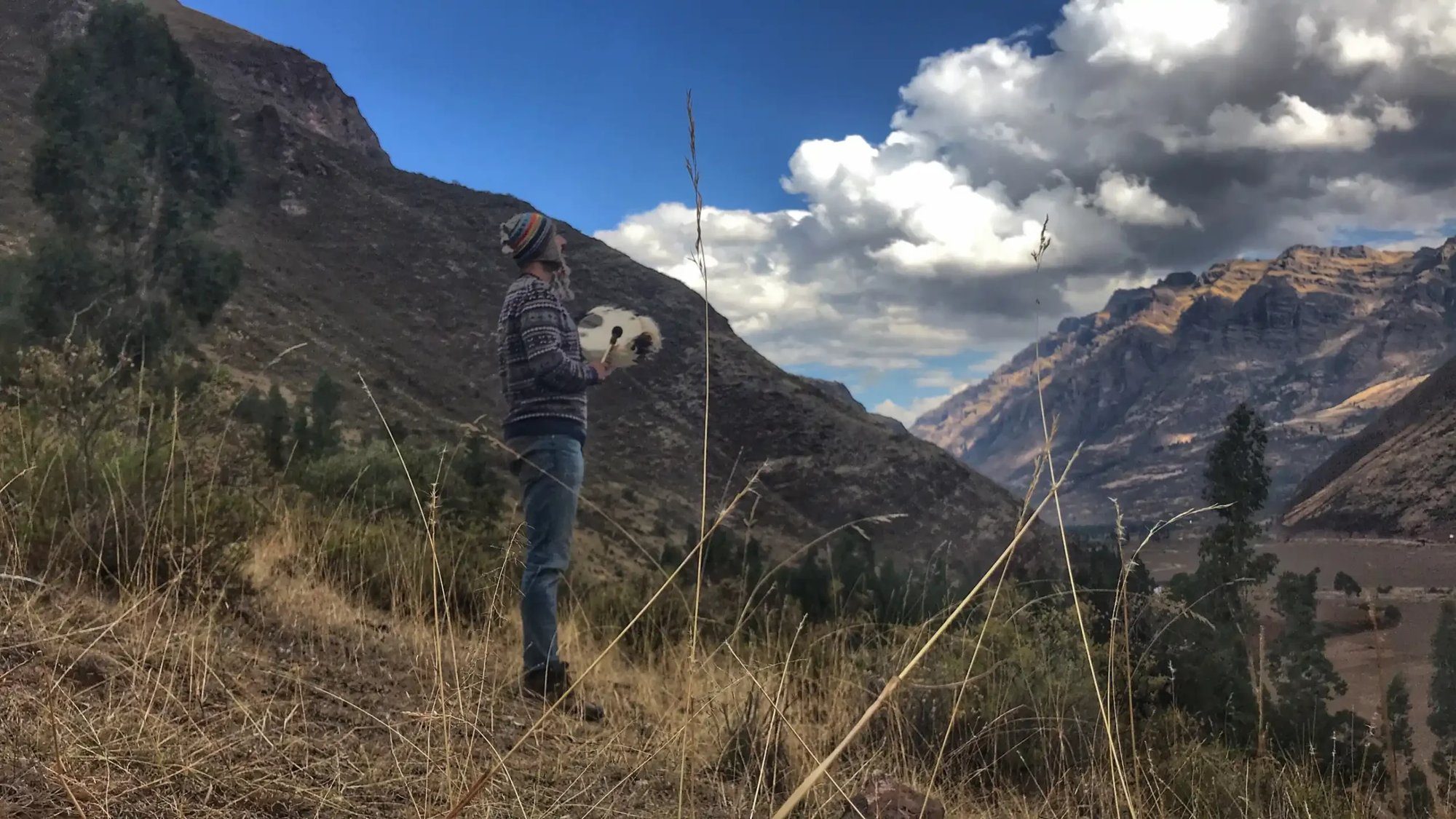 A man playing a flute on a rock above a wide valley, mountains fading into haze behind him.