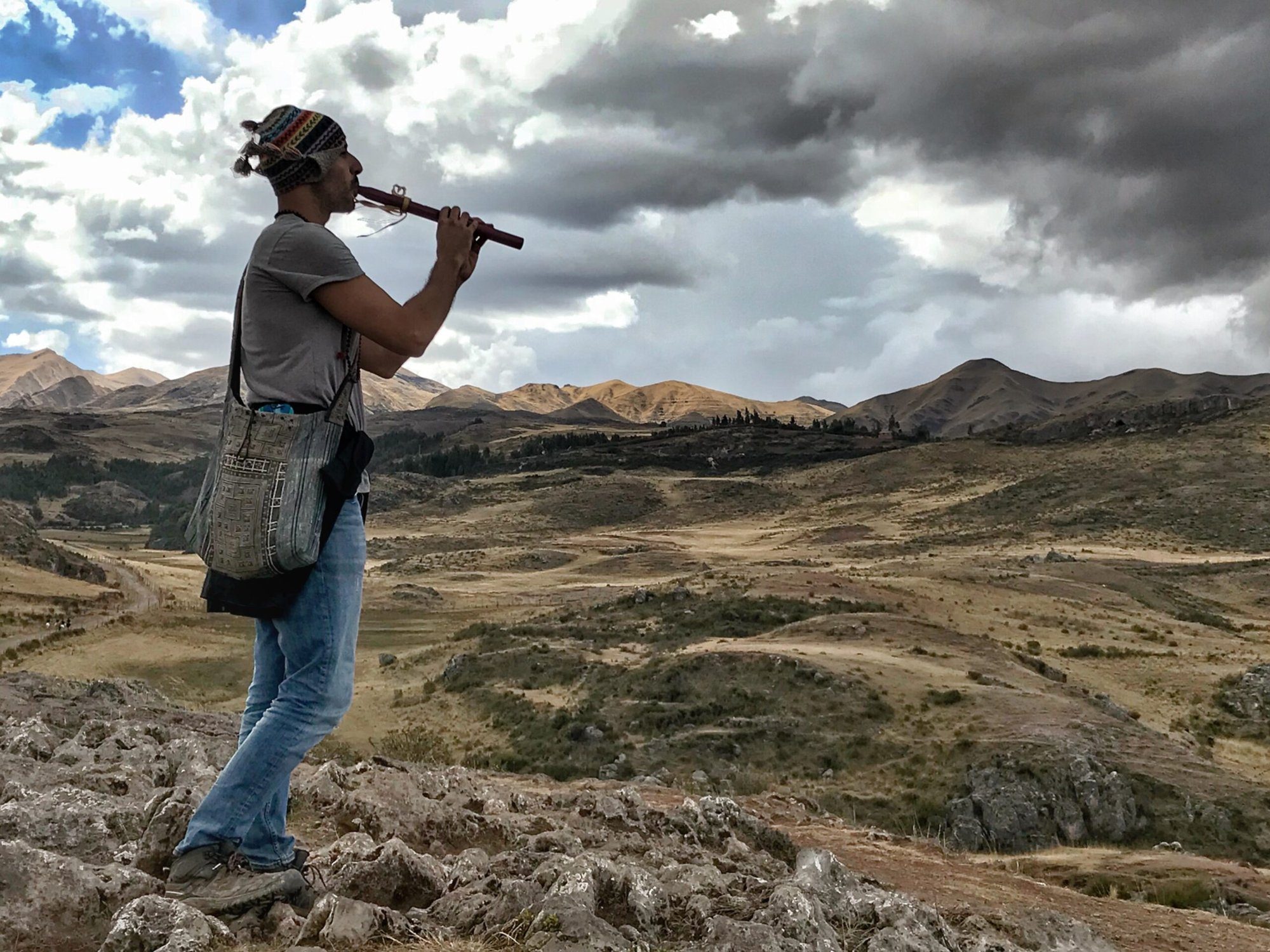 A man playing a flute on a rocky ridge, high golden grassland and a range of bare mountains behind him under a clouded sky.