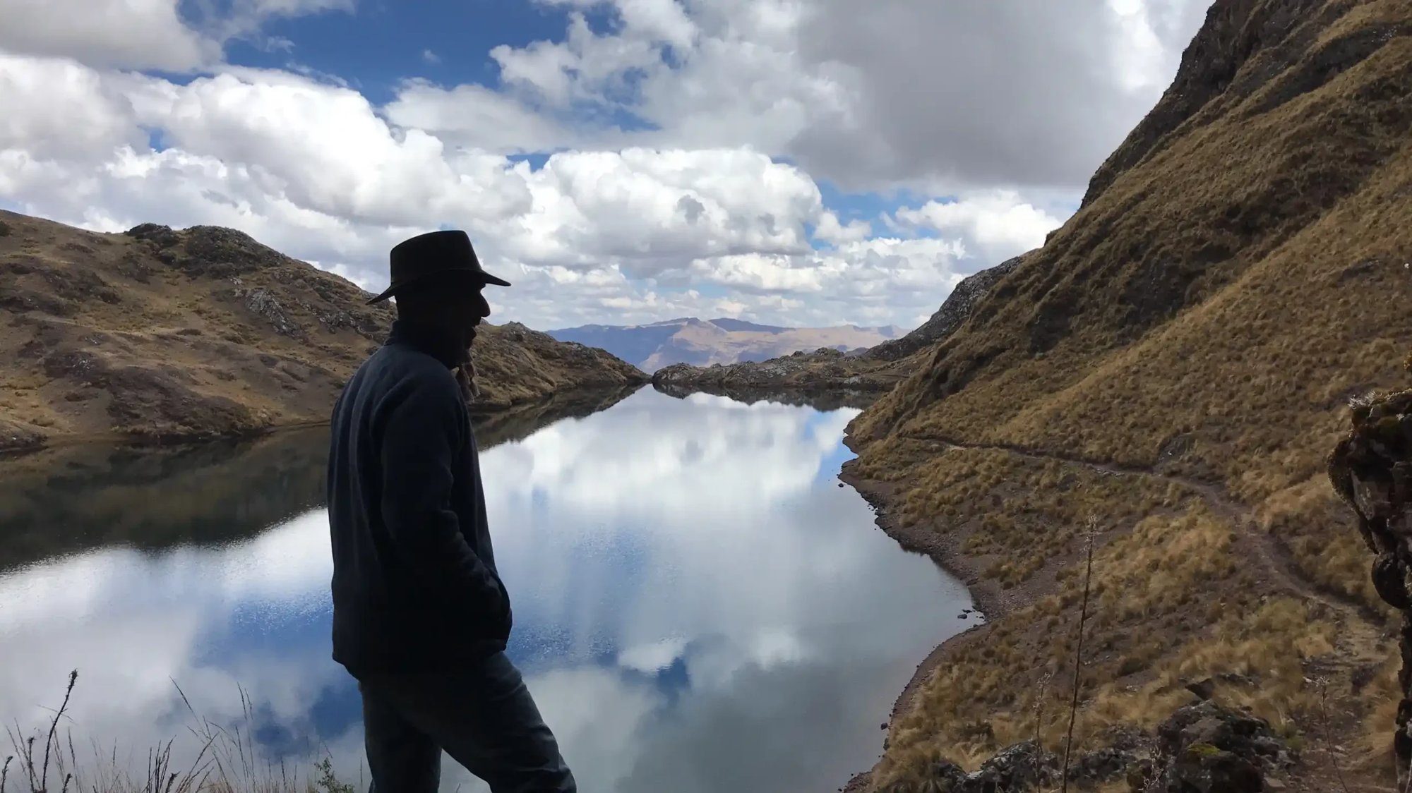 A man in a hat standing at the edge of a mountain lake, his reflection in the still water.