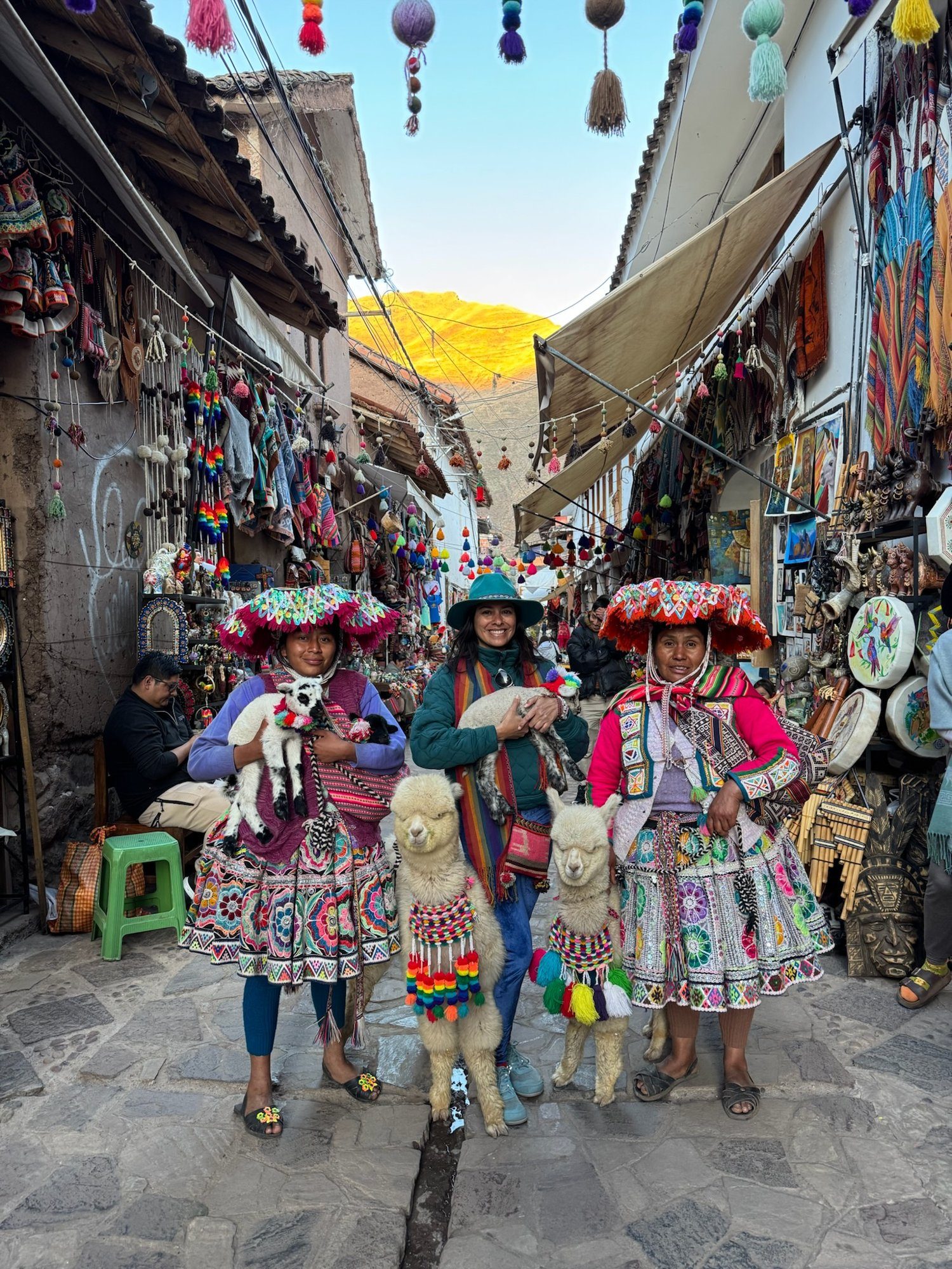 Two women in embroidered hats and skirts and a visitor in a green hat stand with two alpacas in a market street in Pisac, craft stalls on both sides and a sunlit ridge at the end of the lane.