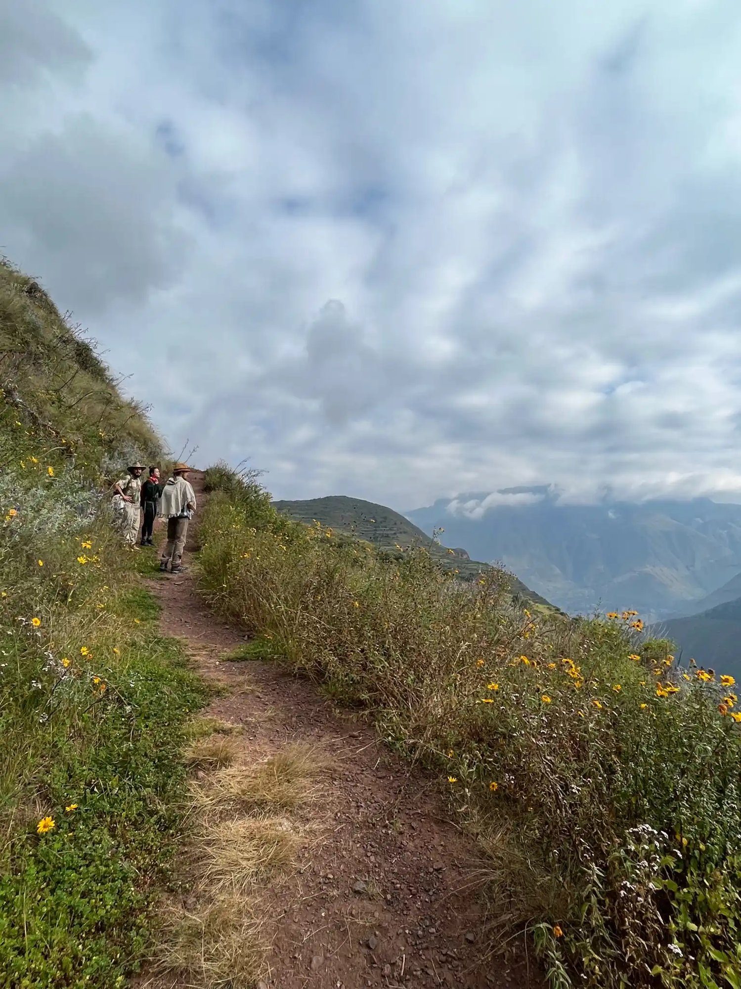 A narrow dirt trail along a green ridge, a walker far ahead and a river valley below.