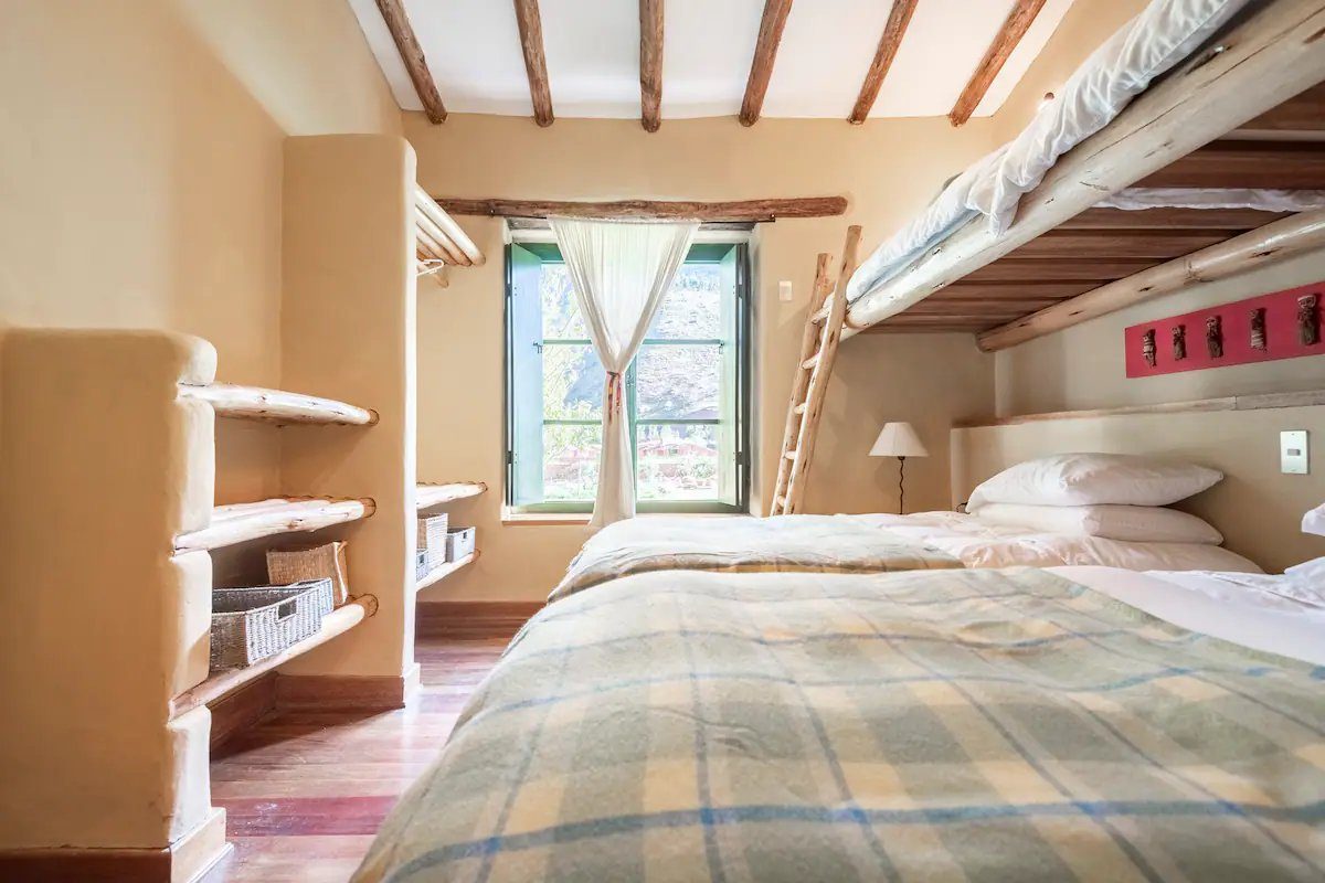 A bedroom with a bunk bed under a beamed ceiling, a checked blanket on the lower bed and a green framed window.