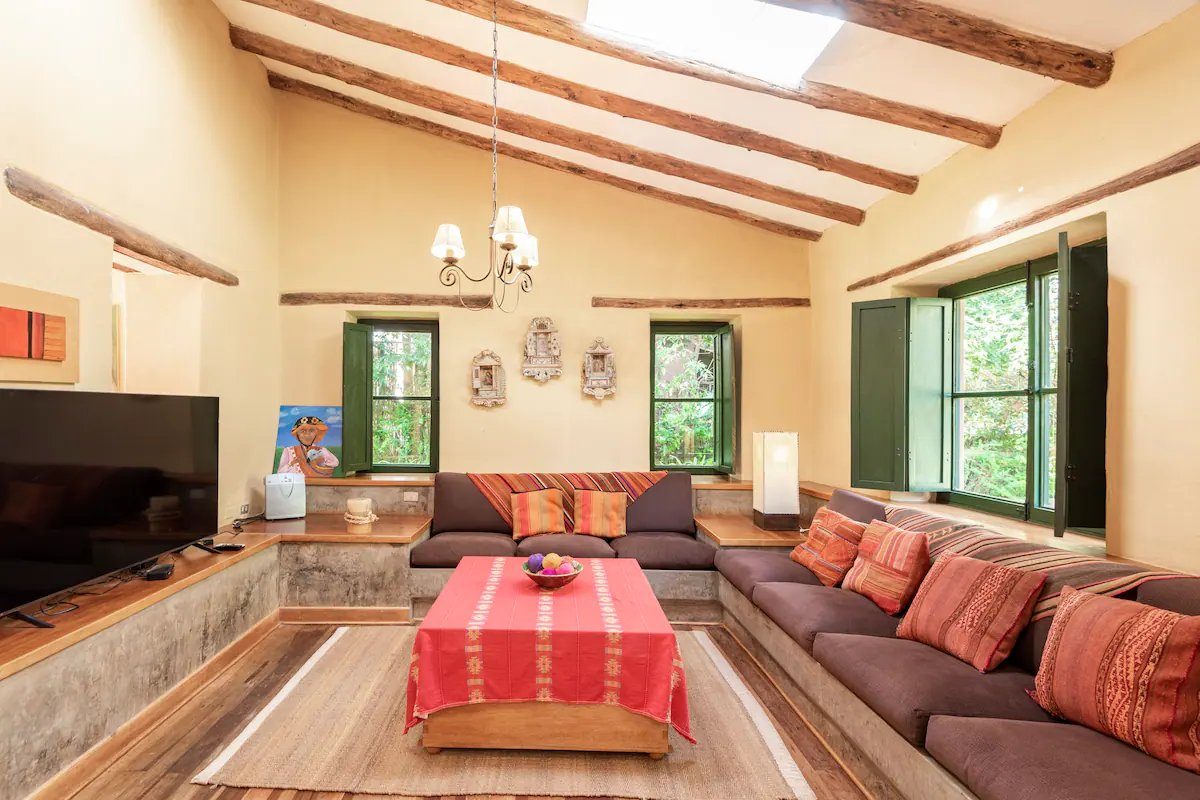 A sitting room with a long grey sofa, red and orange cushions, a red woven cloth on the low table and green framed windows.