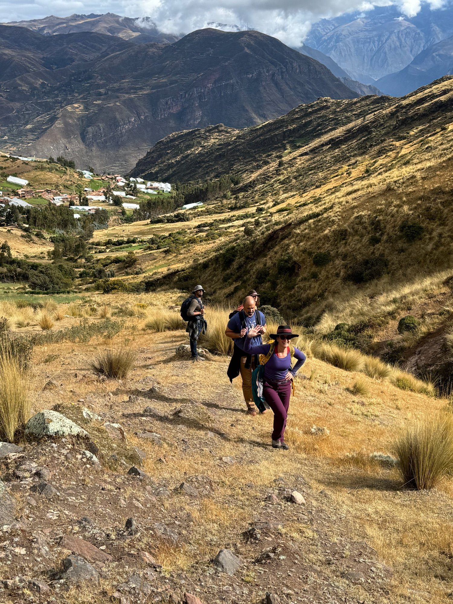 Three people climbing a grassy ridge high above a valley village, one turning back toward the camera with a hand raised.
